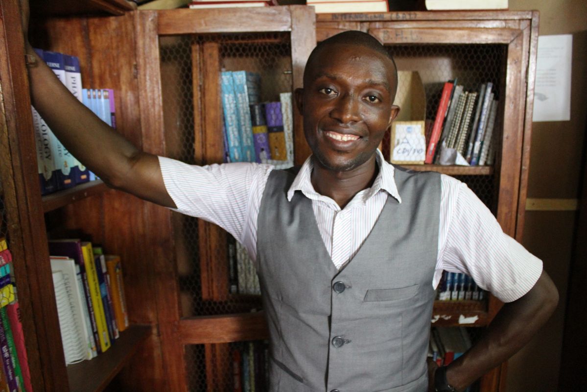 Portrait of Alex Bo standing with his arm extended and resting on a bookshelf, his other hand on his hip. He is smiling at camera.