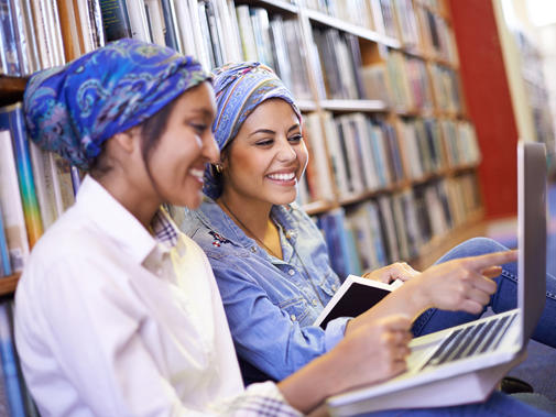 Two women smiling and sat on the floor leaning against a bookshelf with laptops open