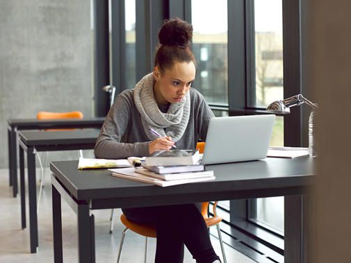A woman sat alone at a library table near a window in front of books and laptop. She is concentrating on her screen with a pen in hand.