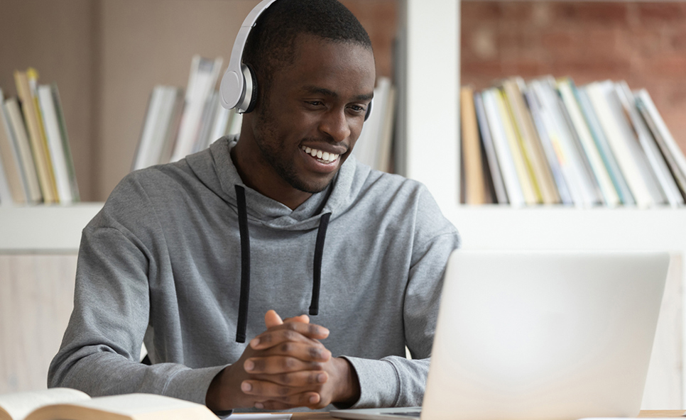 A man wearing headphones at his laptop. He is smiling at the person he is conversing with on his screen. A bookshelf is out of focus in the background.
