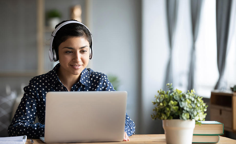 A woman in headphones sits at a desk in the library and smiles at her open laptop