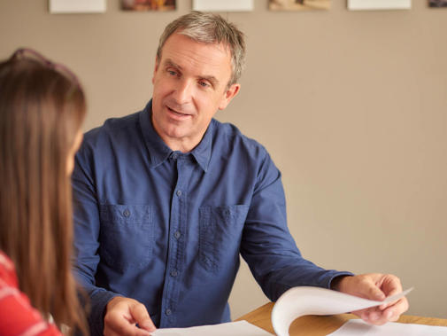 A man sits speaking to a woman with her back to the camera. He is holding open a stapled document.