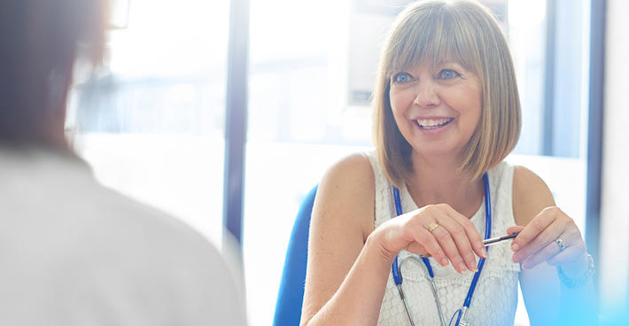 A woman wearing a stethoscope and holding a pen smiles and speaks to a second person who is blurred and not facing the camera