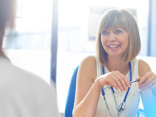 A woman wearing a stethoscope and holding a pen smiles and speaks to a second person who is blurred and not facing the camera