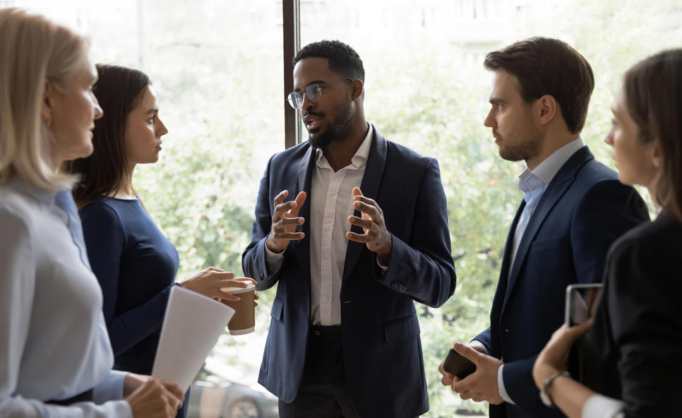 People in business attire stood in a tight circle in front of a window. A man is explaining something and gesticulating with his hands