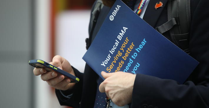 Man at ARM studies phone whilst holding a BMA benefits booklet in his other hand. He wears a BMA rainbow lanyard.