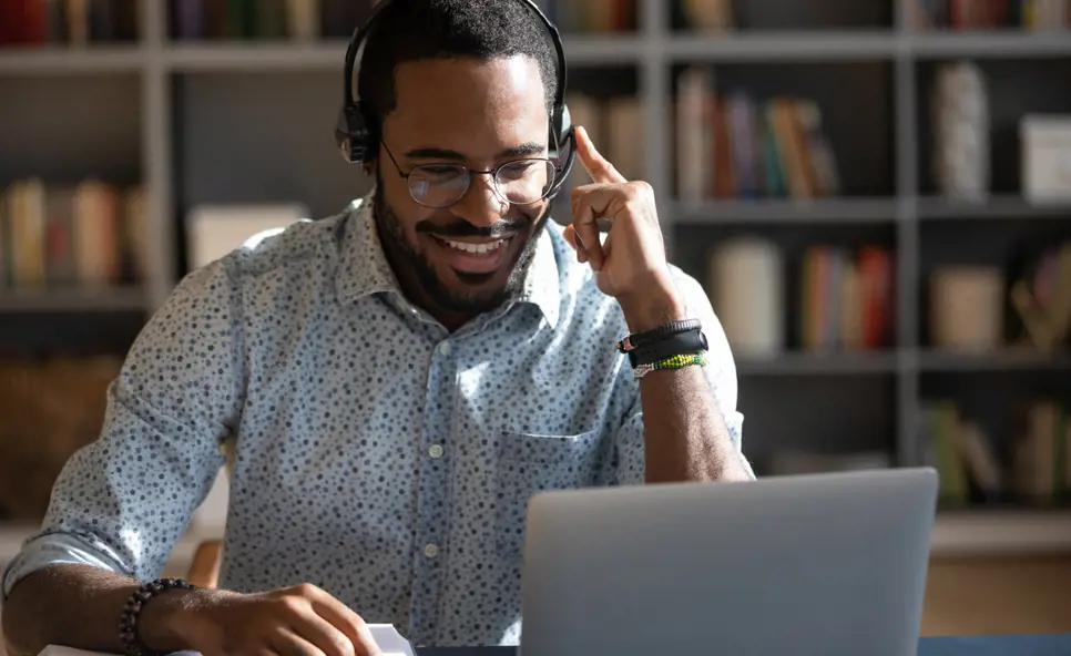 A smiling man wearing a headset in conversation at his laptop. A metal framed bookshelf is in the background and out of focus.